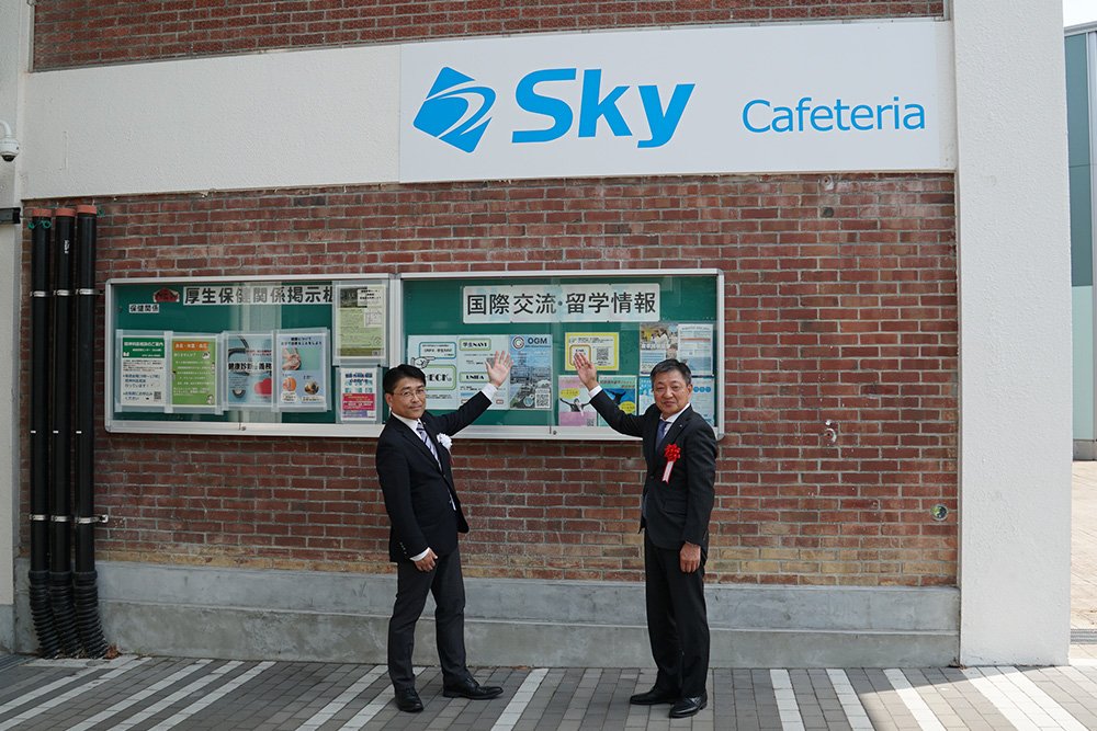 写真：看板の前に立つ松本副学長と寺下執行役員
